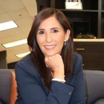 Yissel Munoz, a woman with shoulder-length dark brown hair, wearing a navy-blue blazer, sits in a modern office space, smiling at the camera.